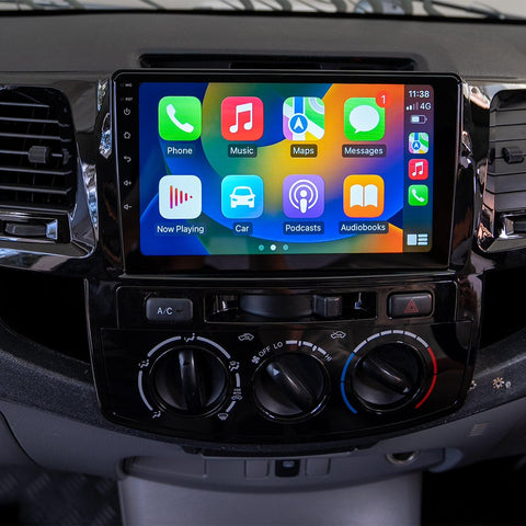 Dashboard of the Toyota Hilux 2005-2015 featuring a multimedia system with Apple CarPlay. The screen displays app icons for Phone, Maps, Messages, Now Playing, Car, Calendar, Settings, and Google Maps. Below the screen are controls for air conditioning and other car functions. The interface indicates a connected iPhone with 4G signal and time display. Type 1.